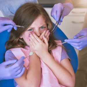 Little girl in dentist chair covering mouth, while gloved hands stick dental tools at her, representing dental anxiety