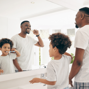 Dad and son brush their teeth in front of bathroom mirror, representing teeth cleaning
